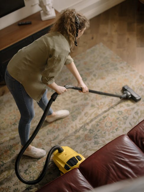 A person cleaning a patterned carpet with a yellow and black vacuum cleaner, holding a cloth to the vacuum's dust container. The image shows the carpet's surface with intricate floral and geometric de