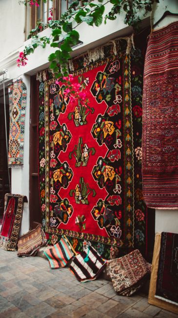 An outdoor shopfront displaying a collection of traditional and patterned woven rugs and carpets arranged on the pavement and leaning against the building facade. The shop has an open entrance with te